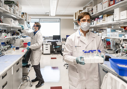 Two people working in laboratory. One carries a tray of test tubes.